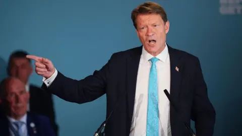PA Reform UK deputy leader Richard Tice speaks during a press conference at the Royal Horseguards Hotel in London. He is pointing with his finger to the left. He is wearing a black suit jacket, white shirt and a blue tie. He has short brown hair.