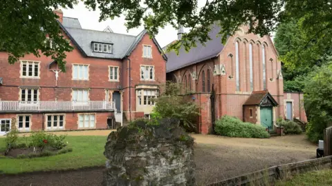 BBC A view of two large redbrick Victorian buildings with smart gardens in the foreground. Both buildings are grand at least 3 stories high.