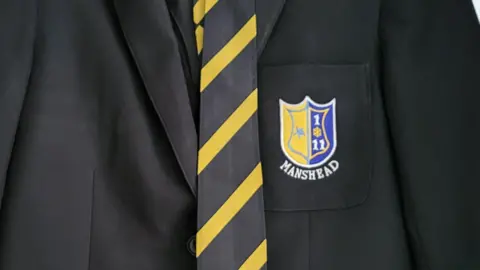 Kerry Roblett A black school blazer with a black-and-yellow tie hanging down the middle. On the pocket of the left breast is a blue and yellow shield and the word "Manshead" underneath