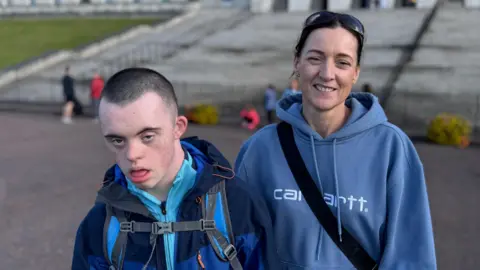 BBC Suzy Ward and her son Eoyn stand in front of a large cream building with a stone staircase leading up to it. The boy has a brown buzz-cut. He wears a light blue top with a navy coat on over it. He has a backpack with blue straps. The woman, who stands to the boys left wears a navy hoodie. She has a black bag strap across her chest. 