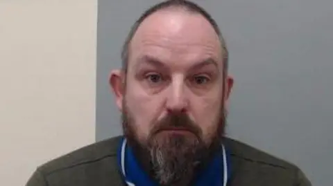 PSNI A man with a bald head and grey beard and dark eyes. He has a blue collar on and grey top. He is looking at the camera and behind him is a white and blue wall.