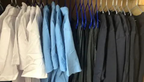 Getty Images A rail of uniforms with white shirts, blue shirts, grey trousers and black or grey sweatshirts on hangers.