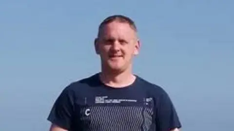 PSNI A blond young man with blue eyes wearing a dark blue top. He is smiling and standing up against a blue sky.