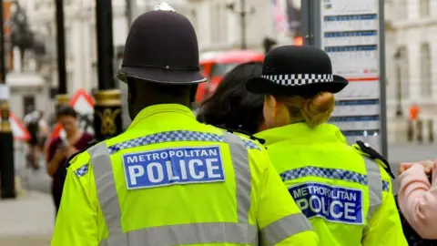 Getty Images Two police officers, a man and a woman, seen from behind in a busy street walking past a bus stop. Their uniform reads METROPOLITAN POLICE.