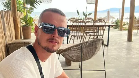Family photo Nathan Osman is looking at the camera while wearing a white T-shirt and large, square black sunglasses. A black bag strap can be seen over his right shoulder and his has very short, light brown hair and a trimmed goatee. In the background is an outdoor bar or cafe and the sea and coastline can be seen in the distance