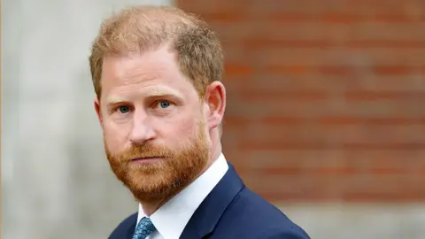 Getty Images Prince Harry is wearing a dark suit jacket, white dress shirt, and blue patterned tie. The background consists of a brick wall on the right side and a light-colored concrete structure on the left side.