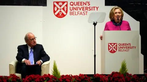 Getty Images George Mitchell sitting on a cream leather chair on a stage, he is wearing a navy suit with blue tie and white shirt, he has white hair and glasses. He is looking to the left where Hillary Clinton is standing at a lectern on the stage talking into a microphone. She is wearing a pink top. She has blonde hair.