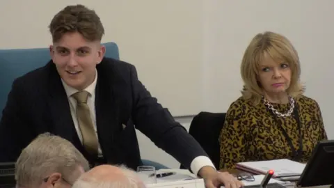 Warwickshire County Council Warwickshire County Council leader George Finch sat next to council chief executive Monica Fogarty. He is wearing a dark striped suit, white shirt and gold tie. She is wearing an animal print blouse and thick chain necklace.