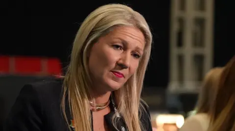 BBC Laura Anne Jones wearing a black top, in conversation with a person slightly off camera. She has a black backdrop behind her and is wearing a Reform party lanyard.