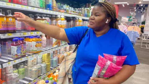 Danielle Kaye/BBC Yanique Clarke, a nursing student in Manhattan who identifies as lower-income, shops in the grocery section of a Target location on 10 Sept. 2025. She is holding two bags of produce reaches out to grab a drink from a grocery aisle.