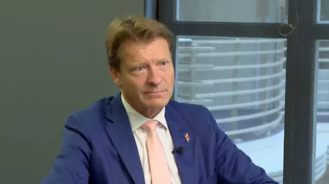 BBC Richard Tice, Deputy Leader of Reform UK, sitting in the BBC Millbank offices during an interview with BBC South East. Looking off camera, wearing a blue suit with a Union Jack pin and a pink tie