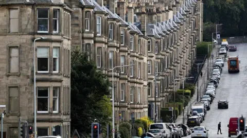 PA A row of tenement flats on a hill with a line of parked cars outside. A lone man can be seen crossing the road