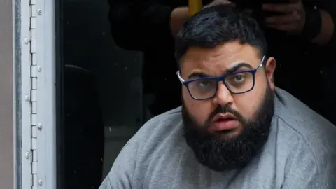 Athena Abubakr Khawar emerging from security truck in grey jumper. He has a black beard and black short hair and wears glasses