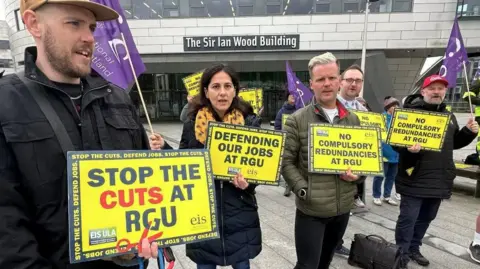 BBC A group of staff at Robert Gordon University striking in Aberdeen. They are holding yellow placards which say "stop the cuts at RGU" and "defending our jobs at RGU". They are stadning in front of a building which has "The Sir Ian Wood Building" above the door.