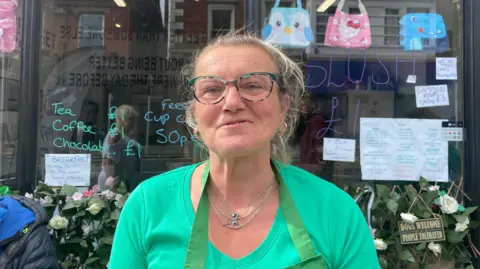 BBC Sam Elliott has blonde-brown hair tied back in a ponytail. She is wearing a green t-shirt green apron and green glasses and smiling. She's standing in front of a cafe window. Menu items and prices are displayed on the window in colourful writing. 