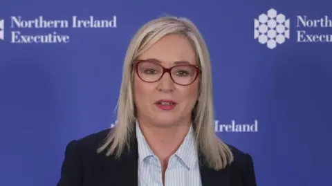BBC Michelle ONeill in a blue blazer and striped blue top. She stands at a mic. She has blonde hair and glasses.