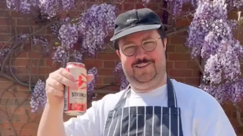 Al Nash Al Nash, a white man with a moustache and glasses, holds a can of Strongbow cider. He is wearing a chef's apron and a cap because he is at a barbecue. He is standing against a red brick wall with wisteria growing over it.