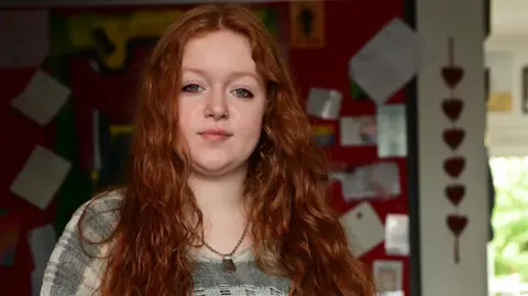 Equality Commission Rosie is looking into the camera. She has long curly auburn hair. She's in a classroom and there is school work displayed on the walls. She's wearing a grey stripped jumper and a silver necklace. 