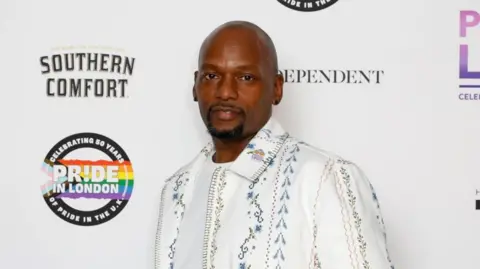 Getty Images A man poses against a Pride in London backdrop at a red carpet event. The photo, which shows him from the chest up, shows him wearing a white jacket with floral embroiding. He has a goatee and isn't smiling at the camera.