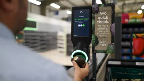 Tesco A man in a blue shirt holds an avocado to one of Tesco's new avocado scanners, the machine glows green and a screen reads 'ripe and ready to eat'