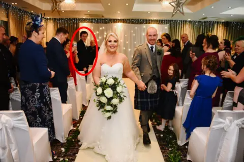Belvedere Images A happy bride and groom walking back up the aisle after their wedding, with beaming smiles on their faces. Many of the guests are applauding them. Andrew Hillhouse can be seen in the background as one of the attendees, with a red ring overlain on the image to draw the eye to him.
