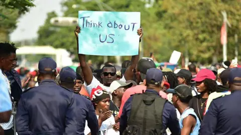 AFP via Getty Images A protester holds a placard that says "think about us" in English as demonstrators gather in Luanda. Some policemen with their backs to the camera can be seen in the foregound.