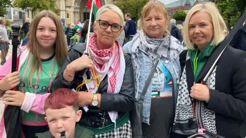 BBC Mary Kelly and family members at the Belfast protest on Saturday.  Three women with blonde hair, a young girl with long blond hair and a young boy with short red hair stand in front of St Anne's Cathedral.  The woman in the middle is  carrying a Palestinian flag. The boy is blowing into a whistle.