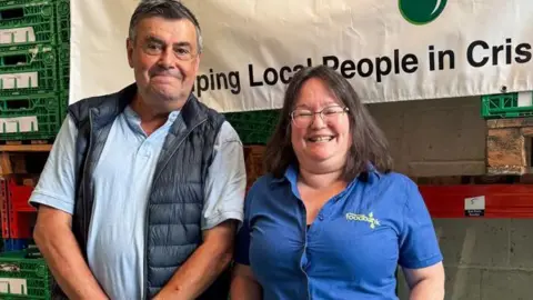 BBC Mike and Maria smile at the camera while standing in a warehouse, part of a foodbank banner behind them. Mike is wearing a blue polo top and dark gilet while Maria wears a blue shirt with foodbank branding.