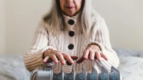 Getty Images An older woman wearing a thick cream cardigan sits on a bed holding her hands over an electric radiator.