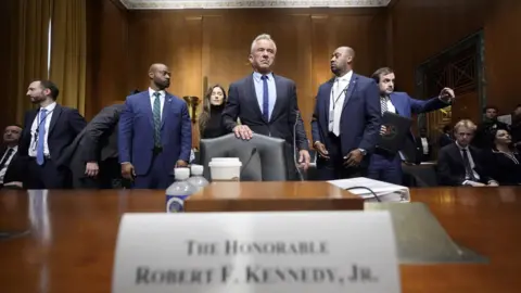 AP Robert F Kennedy Jr stands behind a chair, pushed up to a wide, wooden table. He is wearing a smart, tailored, dark suit. He is flanked by other similarly dressed men. On the table is a sign with his name on it, some plastic bottles and a white disposable drinks cup.