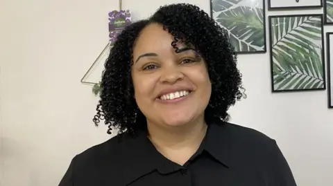 BBC A woman with short black, curly hair to her shoulders smiles into the camera. She wears a black shirt and behind her are photos on the wall, with the ones visible showing the fronds of green ferns.