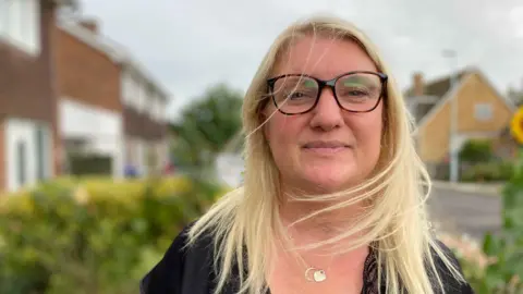 BBC A woman with blond hair and glasses. She is stood in a front garden on a residential street.