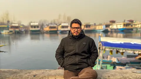 Muneer Speaks/Facebook A man wearing spectacles, a black jacket and brown pants is sitting cross-legged near a lake in Kashmir with houseboats visible in the background.